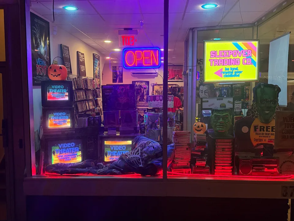 The Sleepover Trading Co. shop window at night under the Community Theater marquee on Main Street in Catskill, NY: a neon OPEN sign, a glowing lightbox, stacked CRT TVs, and Halloween decorations lit in red and purple.