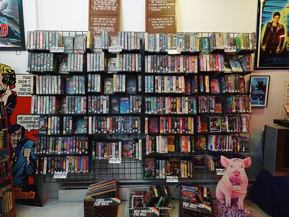 A wall of wire racks packed with VHS tapes for sale upstairs at Sleepover Trading Co. in Catskill, movie posters above, and crates of new arrivals below.