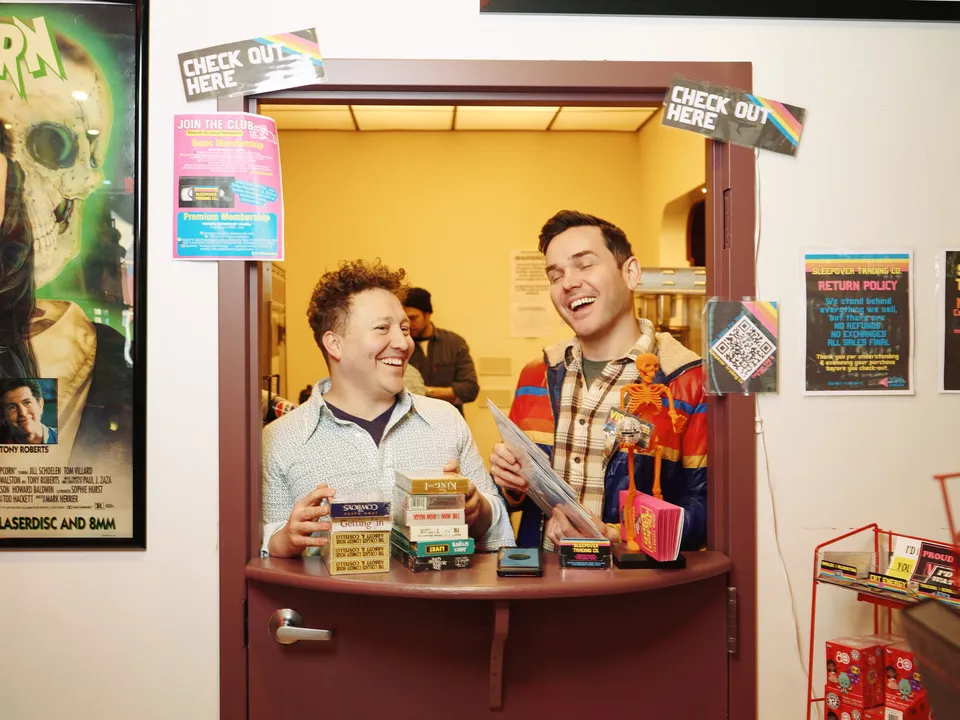 Two people laughing behind the checkout window with a stack of VHS tapes on the counter and Check Out Here signs on the wall.