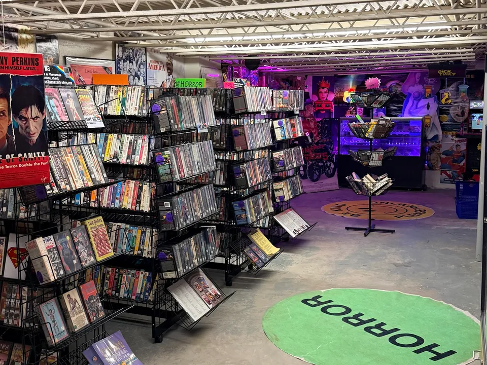 The basement video store from the back: aisles of wire racks full of VHS and DVD rentals, a round green HORROR rug, and the purple neon glow of the arcade corner at the far end.