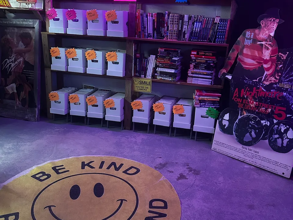 The comic shop corner: shelves of comic longboxes and trade paperbacks, vintage boxed toys along the top, a Freddy Krueger standee, and a round Be Kind Please Rewind rug.